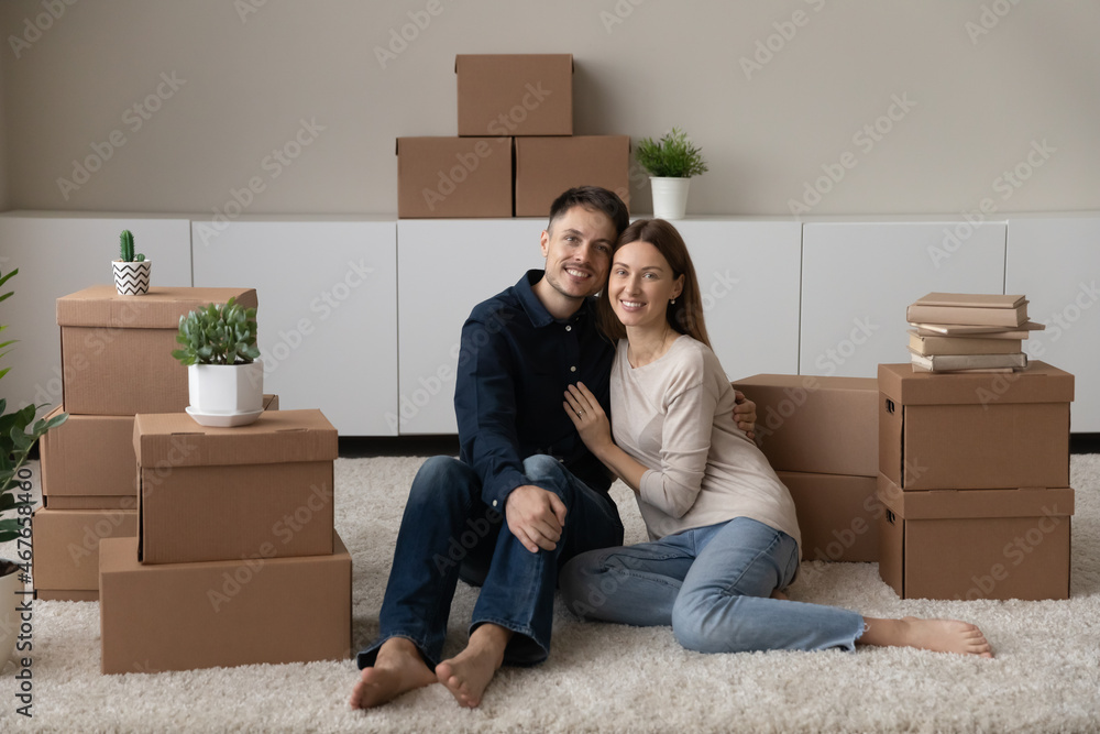 Portrait happy couple hugging, enjoying moving day, sitting on floor with cardboard boxes in new home, smiling woman and man satisfied tenant client looking at camera, relocation and mortgage concept Portrait happy couple hugging, enjoying moving day, sitting on floor with cardboard boxes in new home, smiling woman and man satisfied tenant client looking at camera, relocation and mortgage concept