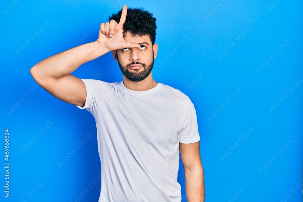 Young arab man with beard wearing casual white t shirt making fun of people with fingers on forehead doing loser gesture mocking and insulting. Young arab man with beard wearing casual white t shirt making fun of people with fingers on forehead doing loser gesture mocking and insulting.