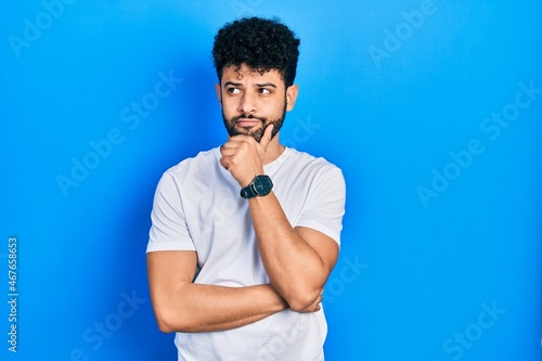 Young arab man with beard wearing casual white t shirt with hand on chin thinking about question, pensive expression. smiling with thoughtful face. doubt concept.