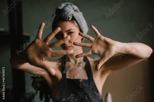 beautiful sculptor girl with a headband and a black apron shows her hands stained with clay. camera focus is on girl's face, blur is on her hands. concept is beauty and art.