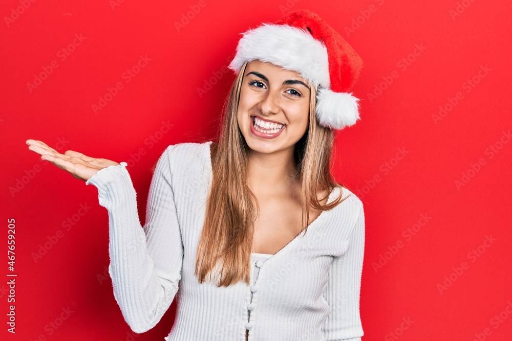 Beautiful hispanic woman wearing christmas hat smiling cheerful presenting and pointing with palm of hand looking at the camera. Beautiful hispanic woman wearing christmas hat smiling cheerful presenting and pointing with palm of hand looking at the camera.