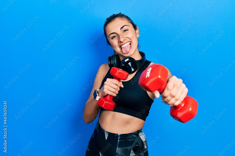 Beautiful hispanic woman wearing sportswear using dumbbells sticking tongue out happy with funny expression. Beautiful hispanic woman wearing sportswear using dumbbells sticking tongue out happy with funny expression.