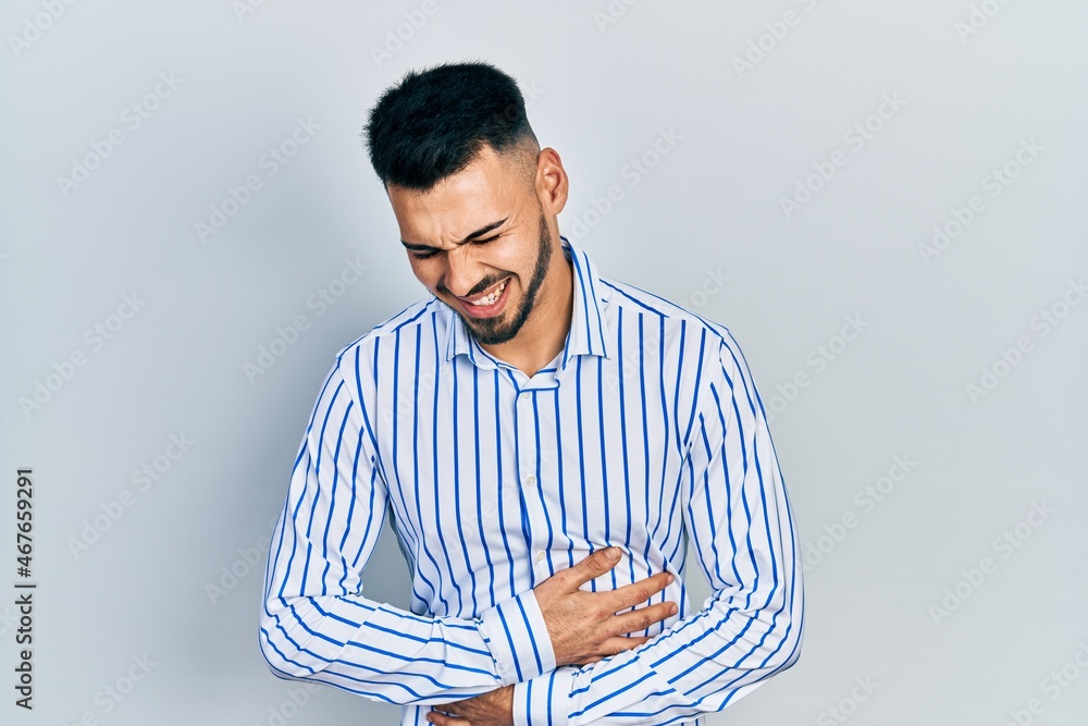 Young hispanic man with beard wearing casual striped shirt with hand on stomach because nausea, painful disease feeling unwell. ache concept. Young hispanic man with beard wearing casual striped shirt with hand on stomach because nausea, painful disease feeling unwell. ache concept.