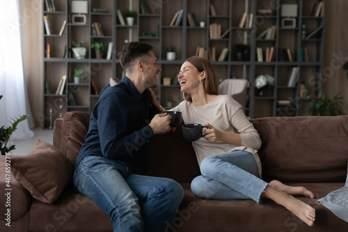 Happy couple chatting, holding cups of tea or coffee, relaxing on cozy couch in living room, smiling young wife and husband laughing at joke together, enjoying romantic date, leisure time at home