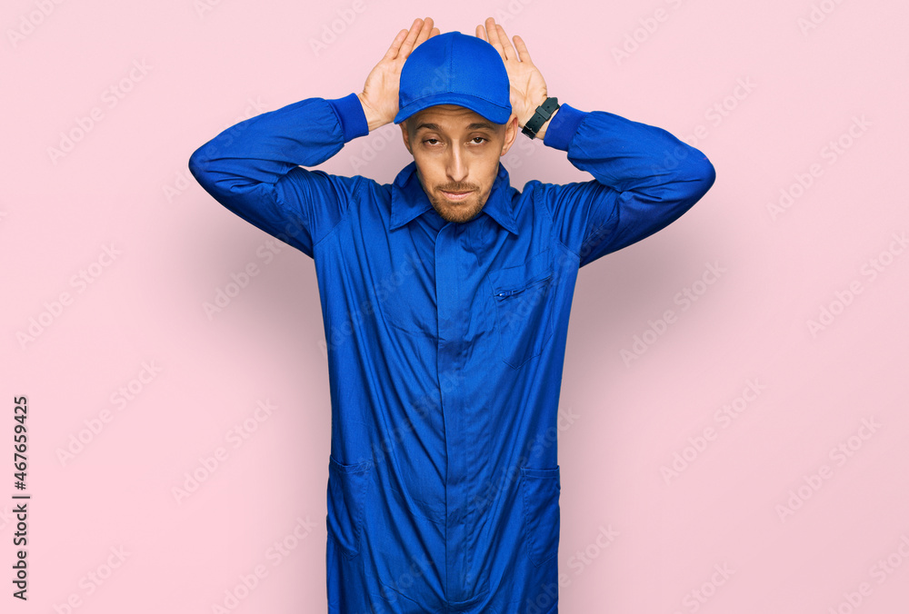Bald man with beard wearing builder jumpsuit uniform doing bunny ears gesture with hands palms looking cynical and skeptical. easter rabbit concept. Bald man with beard wearing builder jumpsuit uniform doing bunny ears gesture with hands palms looking cynical and skeptical. easter rabbit concept.