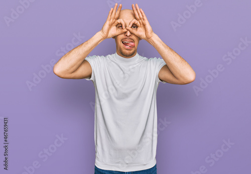 Bald man with beard wearing casual white t shirt doing ok gesture like binoculars sticking tongue out, eyes looking through fingers. crazy expression.
