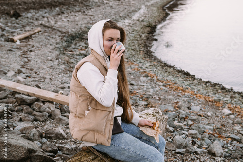 young woman drinking tea on river shore at autumn season