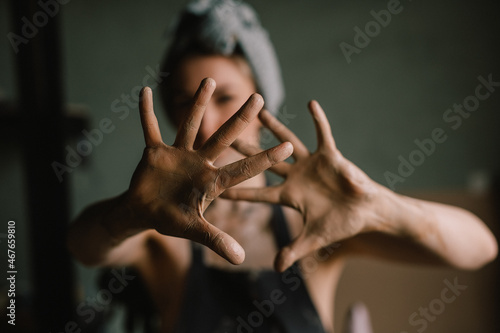 beautiful sculptor girl with a headband and a black apron shows her hands stained with clay. camera focus is on girl's face, blur is on her hands. concept is beauty and art.