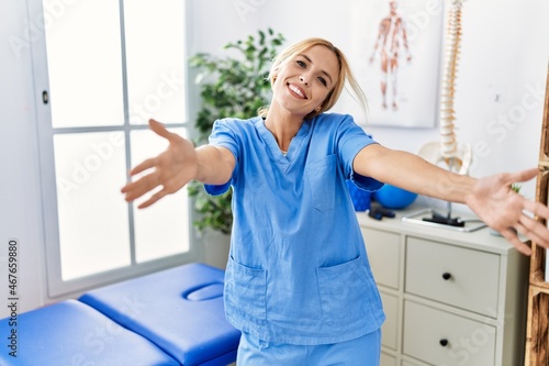 Beautiful blonde physiotherapist woman working at pain recovery clinic looking at the camera smiling with open arms for hug. cheerful expression embracing happiness.