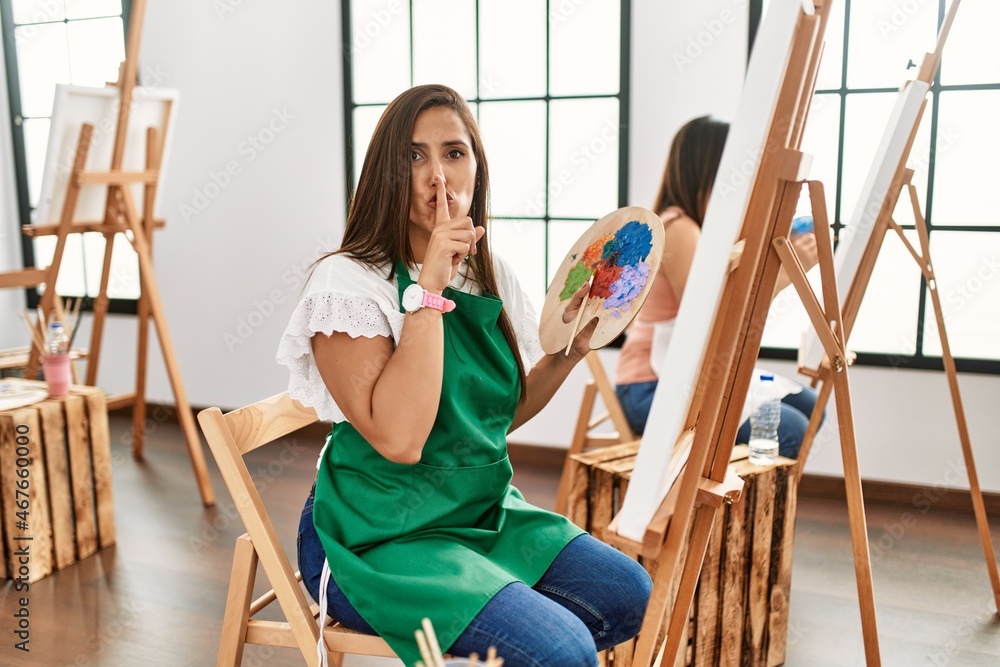 Young hispanic artist women painting on canvas at art studio asking to be quiet with finger on lips. silence and secret concept. Young hispanic artist women painting on canvas at art studio asking to be quiet with finger on lips. silence and secret concept.