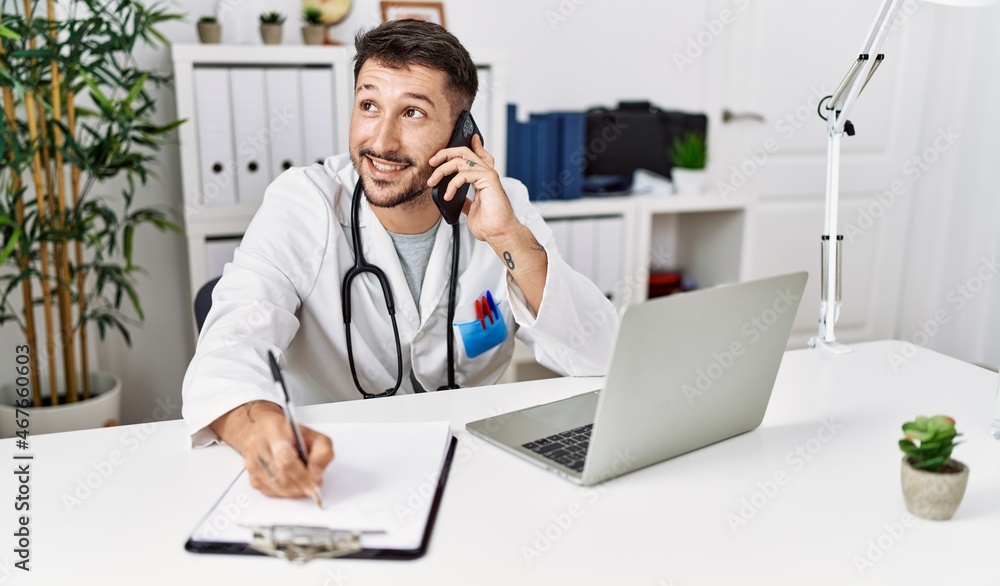 Young hispanic man wearing doctor uniform talking on the smartphone at clinic