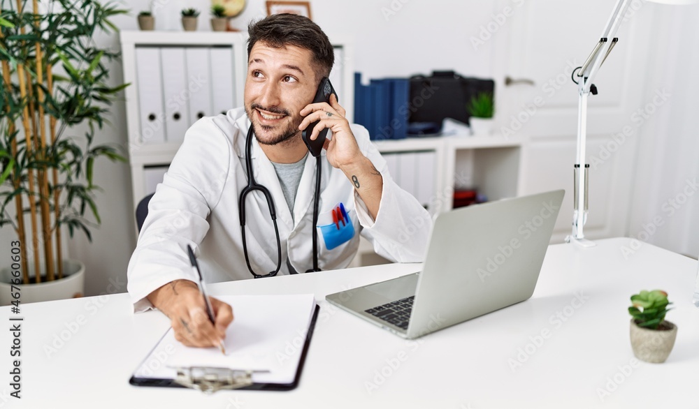 Young hispanic man wearing doctor uniform talking on the smartphone at clinic Young hispanic man wearing doctor uniform talking on the smartphone at clinic