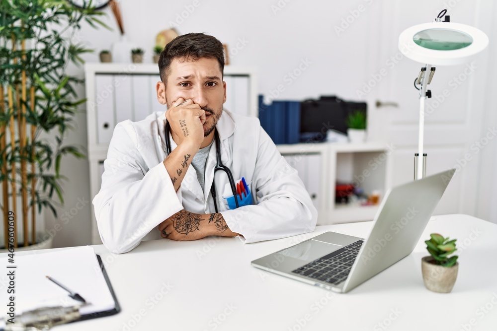 Young doctor working at the clinic using computer laptop looking stressed and nervous with hands on mouth biting nails. anxiety problem.