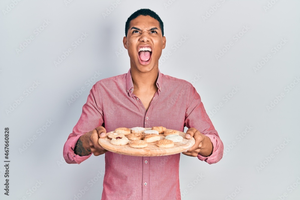 Young african american guy holding tray with cake sweets angry and mad screaming frustrated and furious, shouting with anger looking up. Young african american guy holding tray with cake sweets angry and mad screaming frustrated and furious, shouting with anger looking up.