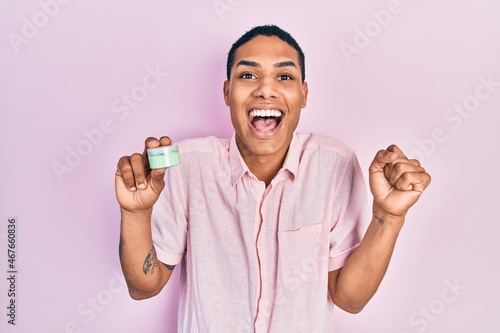 Young african american guy holding face moisturizer cream screaming proud, celebrating victory and success very excited with raised arm © Krakenimages.com