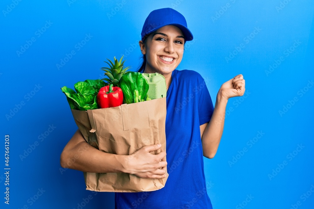 Young hispanic woman wearing courier uniform with groceries from supermarket screaming proud, celebrating victory and success very excited with raised arm Young hispanic woman wearing courier uniform with groceries from supermarket screaming proud, celebrating victory and success very excited with raised arm