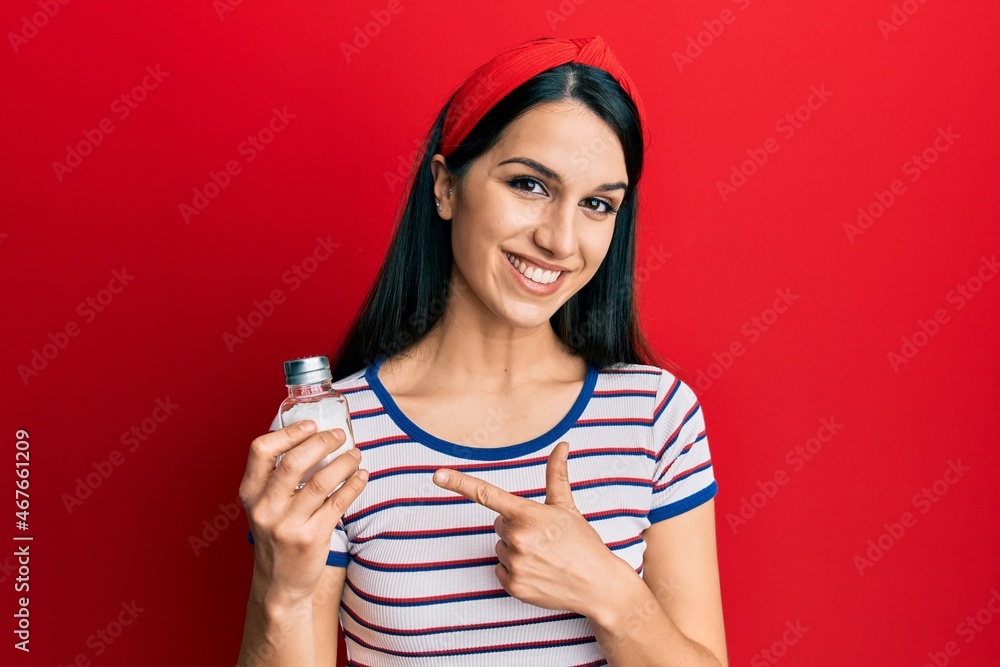 Young hispanic woman holding salt shaker smiling happy pointing with hand and finger Young hispanic woman holding salt shaker smiling happy pointing with hand and finger