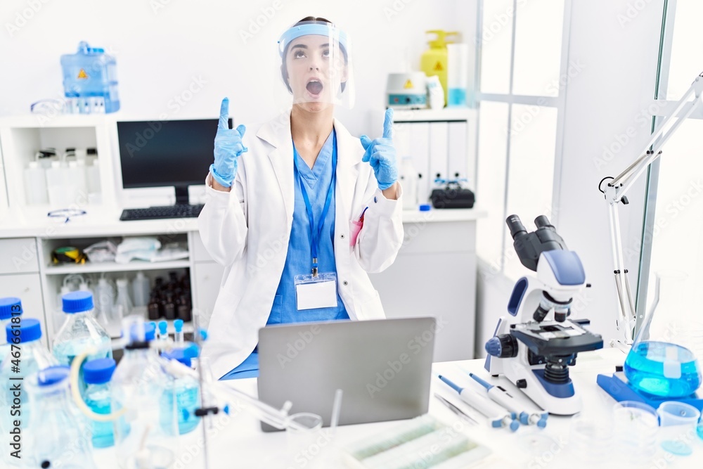 Young hispanic woman wearing scientist uniform amazed and surprised looking up and pointing with fingers and raised arms. Young hispanic woman wearing scientist uniform amazed and surprised looking up and pointing with fingers and raised arms.