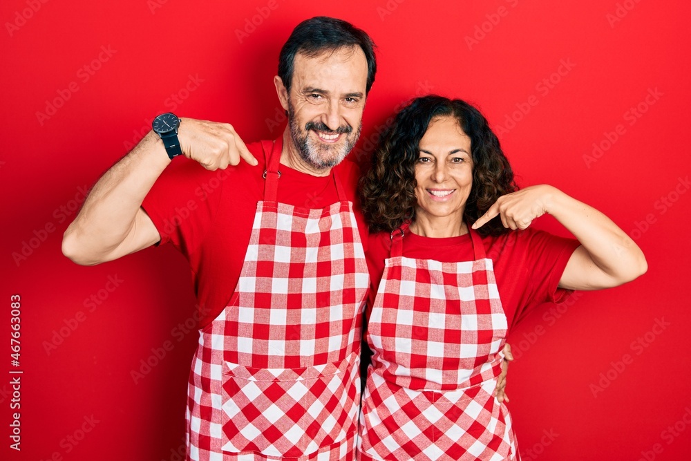 Middle age couple of hispanic woman and man wearing cook apron looking confident with smile on face, pointing oneself with fingers proud and happy. Middle age couple of hispanic woman and man wearing cook apron looking confident with smile on face, pointing oneself with fingers proud and happy.