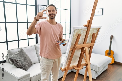 Young hispanic man with beard painting on canvas at home smiling and confident gesturing with hand doing small size sign with fingers looking and the camera. measure concept.