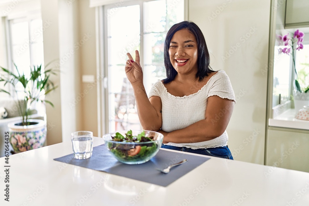 Young hispanic woman eating healthy salad at home smiling with happy face winking at the camera doing victory sign. number two. Young hispanic woman eating healthy salad at home smiling with happy face winking at the camera doing victory sign. number two.