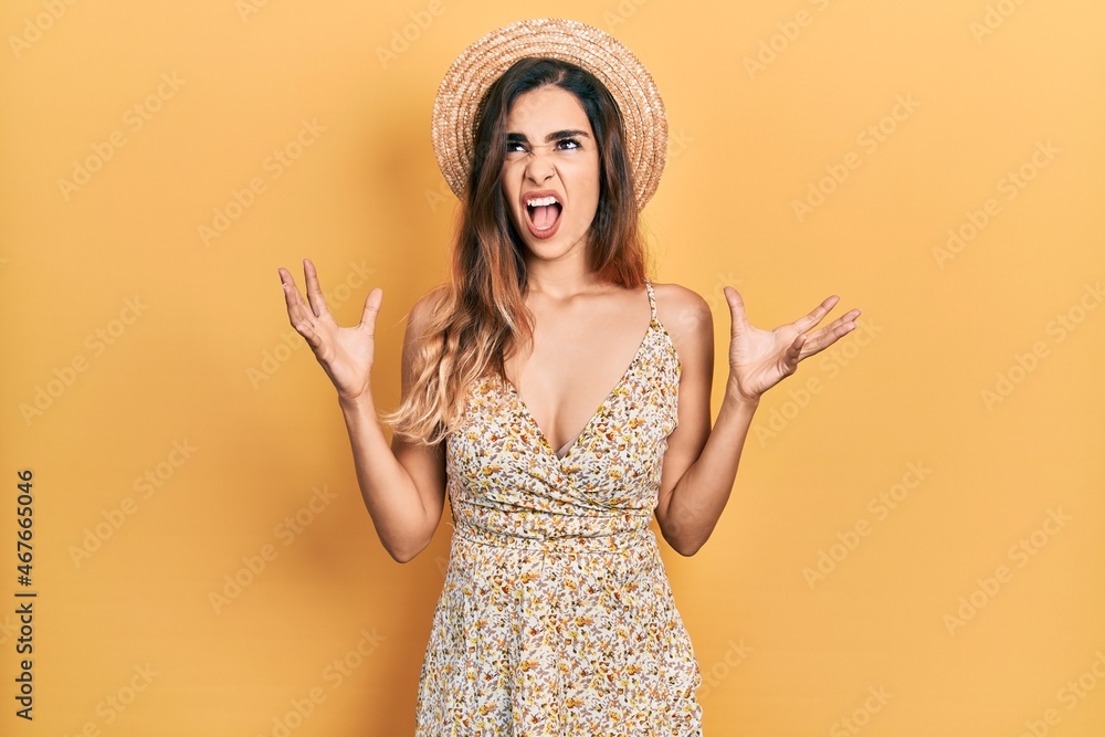 Young hispanic girl wearing summer hat crazy and mad shouting and yelling with aggressive expression and arms raised. frustration concept. Young hispanic girl wearing summer hat crazy and mad shouting and yelling with aggressive expression and arms raised. frustration concept.