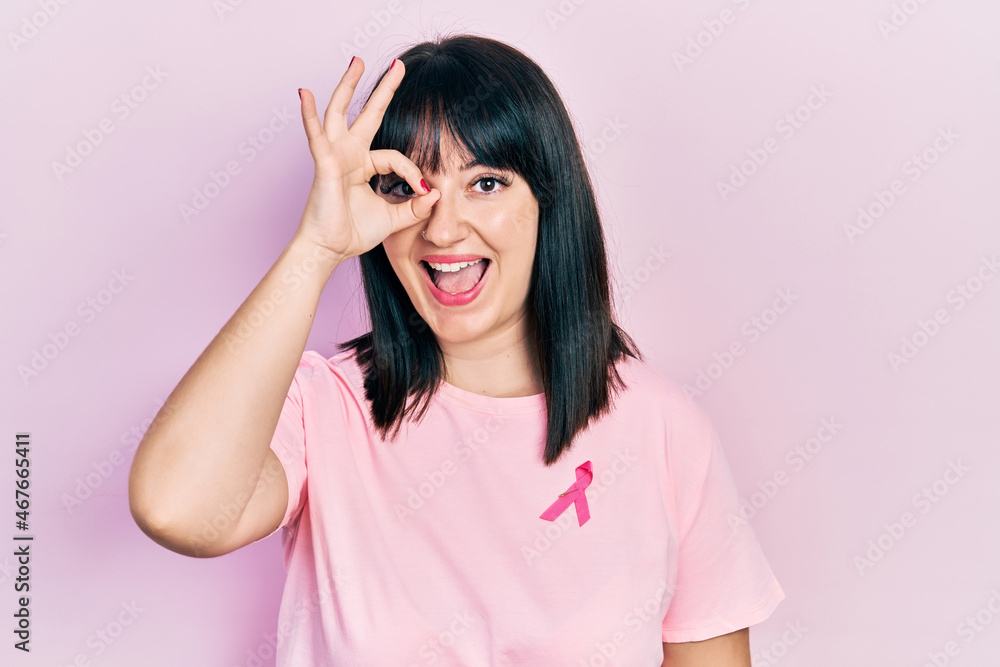 Young hispanic woman wearing pink cancer ribbon on shirt smiling happy doing ok sign with hand on eye looking through fingers Young hispanic woman wearing pink cancer ribbon on shirt smiling happy doing ok sign with hand on eye looking through fingers