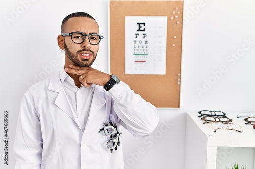 African american optician man standing by eyesight test cutting throat with hand as knife, threaten aggression with furious violence