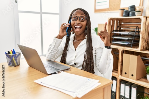 Black woman with braids working at the office speaking on the phone pointing finger up with successful idea. exited and happy. number one.