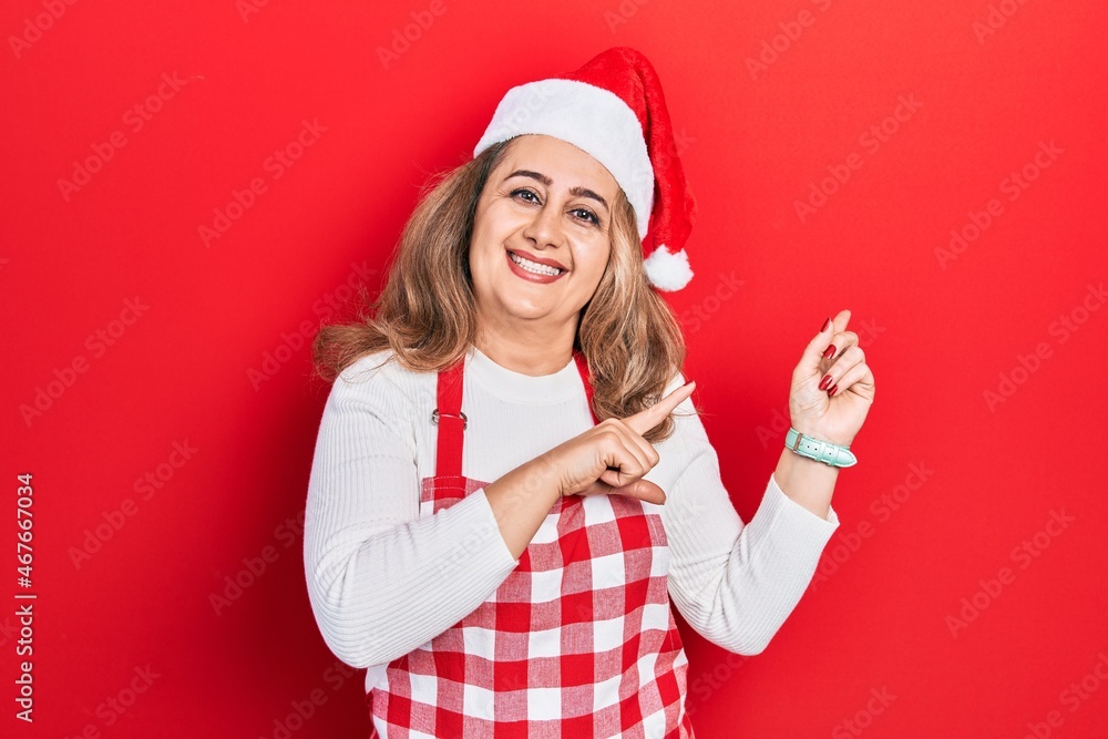 Middle age caucasian woman wearing cook apron and christmas hat smiling and looking at the camera pointing with two hands and fingers to the side. Middle age caucasian woman wearing cook apron and christmas hat smiling and looking at the camera pointing with two hands and fingers to the side.
