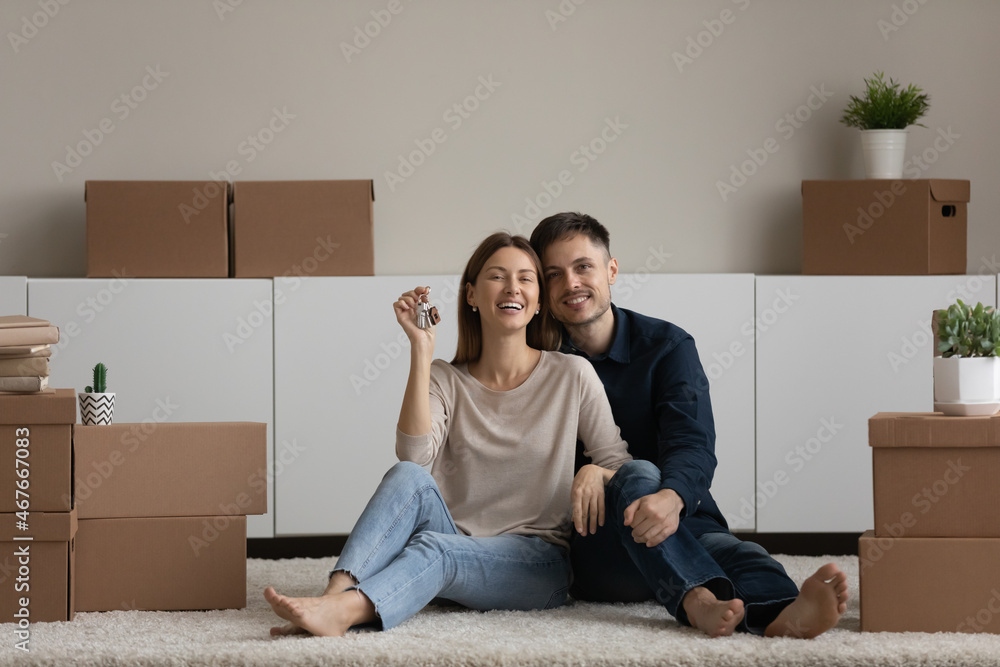 Portrait happy couple demonstrating showing keys, looking at camera, sitting on floor with cardboard boxes in new home, enjoying relocation, smiling woman and man hugging, celebrating moving day Portrait happy couple demonstrating showing keys, looking at camera, sitting on floor with cardboard boxes in new home, enjoying relocation, smiling woman and man hugging, celebrating moving day