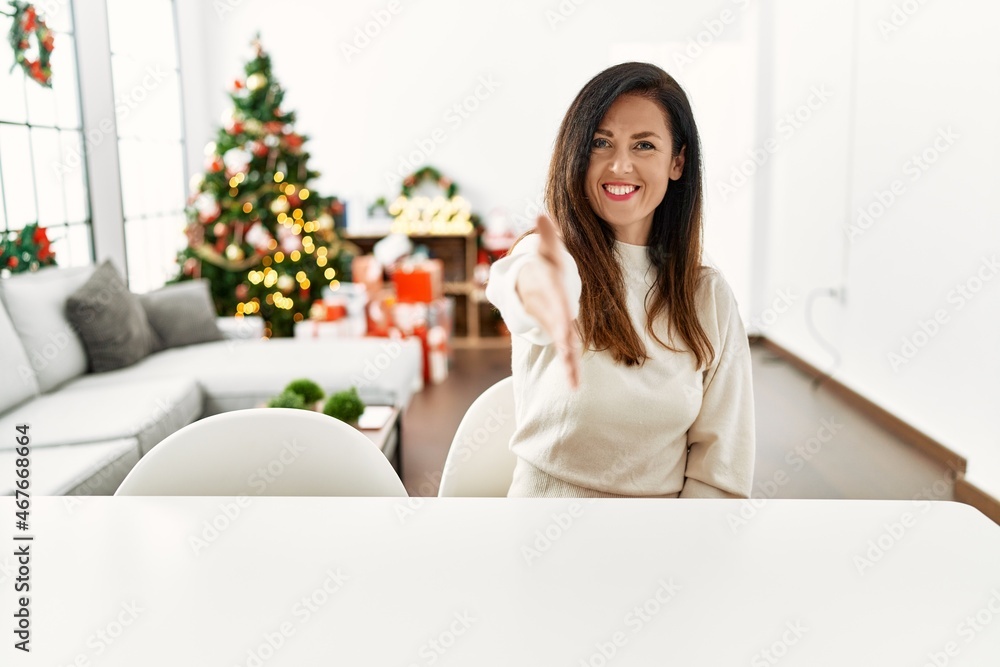 Beautiful caucasian woman sitting on the table by christmas tree smiling friendly offering handshake as greeting and welcoming. successful business. Beautiful caucasian woman sitting on the table by christmas tree smiling friendly offering handshake as greeting and welcoming. successful business.