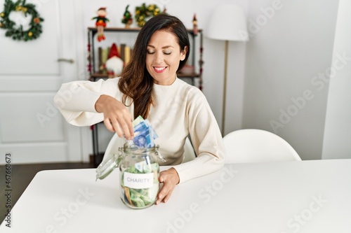 Middle age hispanic woman inserting banknote on charity jar sitting on the table by christmas decor at home