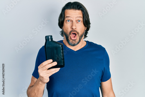 Middle age caucasian man holding motor oil bottle scared and amazed with open mouth for surprise, disbelief face