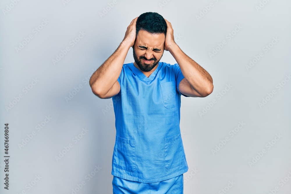 Handsome hispanic man with beard wearing blue male nurse uniform suffering from headache desperate and stressed because pain and migraine. hands on head. Handsome hispanic man with beard wearing blue male nurse uniform suffering from headache desperate and stressed because pain and migraine. hands on head.