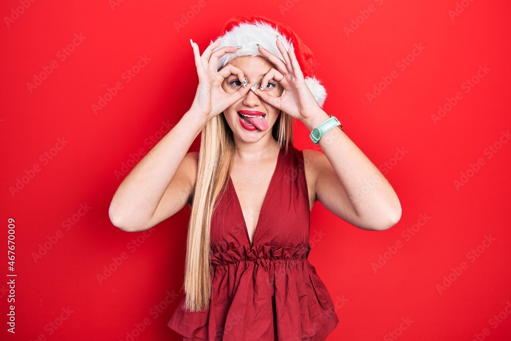 Young blonde girl wearing christmas hat doing ok gesture like binoculars sticking tongue out, eyes looking through fingers. crazy expression. Young blonde girl wearing christmas hat doing ok gesture like binoculars sticking tongue out, eyes looking through fingers. crazy expression.