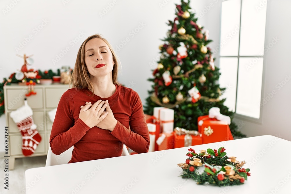 Caucasian young blonde woman sitting on the table by christmas tree smiling with hands on chest with closed eyes and grateful gesture on face. health concept. Caucasian young blonde woman sitting on the table by christmas tree smiling with hands on chest with closed eyes and grateful gesture on face. health concept.