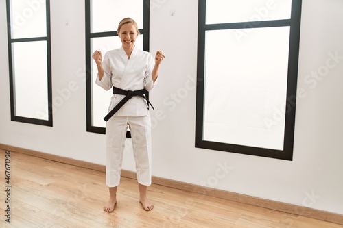 Caucasian young blonde woman wearing karate kimono and black belt screaming proud  celebrating victory and success very excited with raised arms