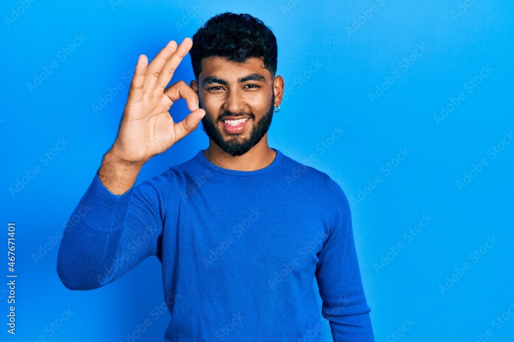 Arab man with beard wearing casual blue sweater smiling positive doing ok sign with hand and fingers. successful expression. Arab man with beard wearing casual blue sweater smiling positive doing ok sign with hand and fingers. successful expression.