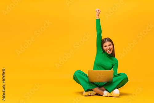 Winner. Excited happy woman sitting on floor with laptop, shouting yes, isolated on yellow