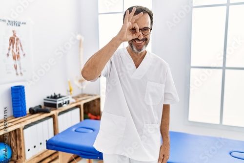 Middle age man with beard working at pain recovery clinic doing ok gesture with hand smiling  eye looking through fingers with happy face.