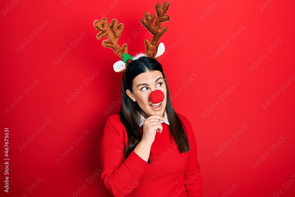 Young hispanic woman wearing deer christmas hat and red nose looking confident at the camera smiling with crossed arms and hand raised on chin. thinking positive.