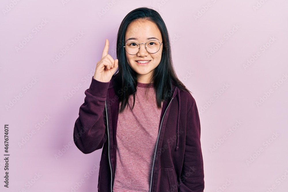 Young chinese girl wearing casual clothes and glasses pointing finger up with successful idea. exited and happy. number one. Young chinese girl wearing casual clothes and glasses pointing finger up with successful idea. exited and happy. number one.