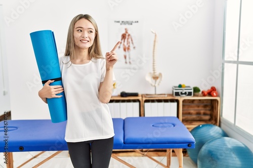 Asian young woman holding yoga mat at pain recovery clinic smiling happy pointing with hand and finger to the side