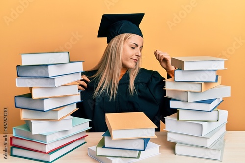 Young caucasian woman wearing graduation ceremony robe sitting on the table stretching back, tired and relaxed, sleepy and yawning for early morning