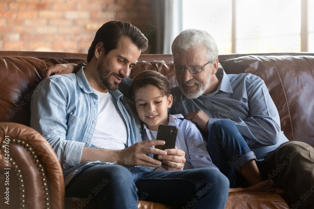 Family day. Three men in family retired grandpa young father preteen boy son grandchild hug on sofa at home relax use modern smartphone. Friendly diverse age male relatives enjoy surfing web on cell