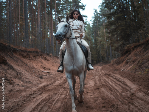 A young beautiful brunette rider in an elegant retro suit riding a white horse in a forest area © Ulia Koltyrina