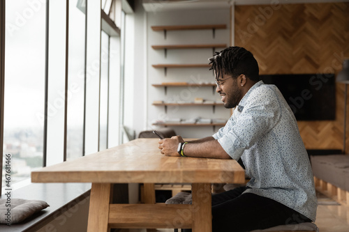 Happy millennial african american biracial man using smartphone, typing message communicating in social network, playing mobile game, shopping in internet store, sitting at table in modern office.