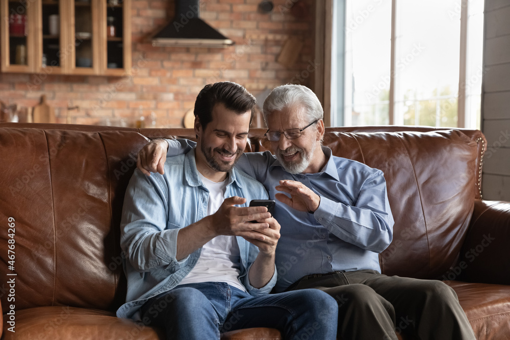 Fototapeta premium Making videocall. Excited retired male talk to grandkid by video call on smartphone with help of grown son. Two happy men of diverse age senior dad adult child engaged in online communication by cell