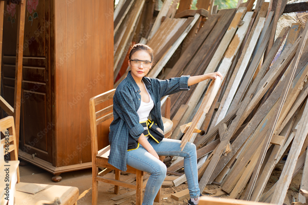 Fototapeta premium Asian female carpenter in blue shirt, jean, tool belt and safety goggles sitting on chair with one hand holding the broken backrest of chair put on her leg while looking at camera in a wood factory.